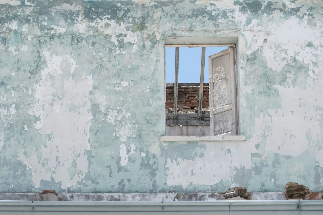 Peeling pale green paint on an old plaster wall around a window with a flaking painted shutter