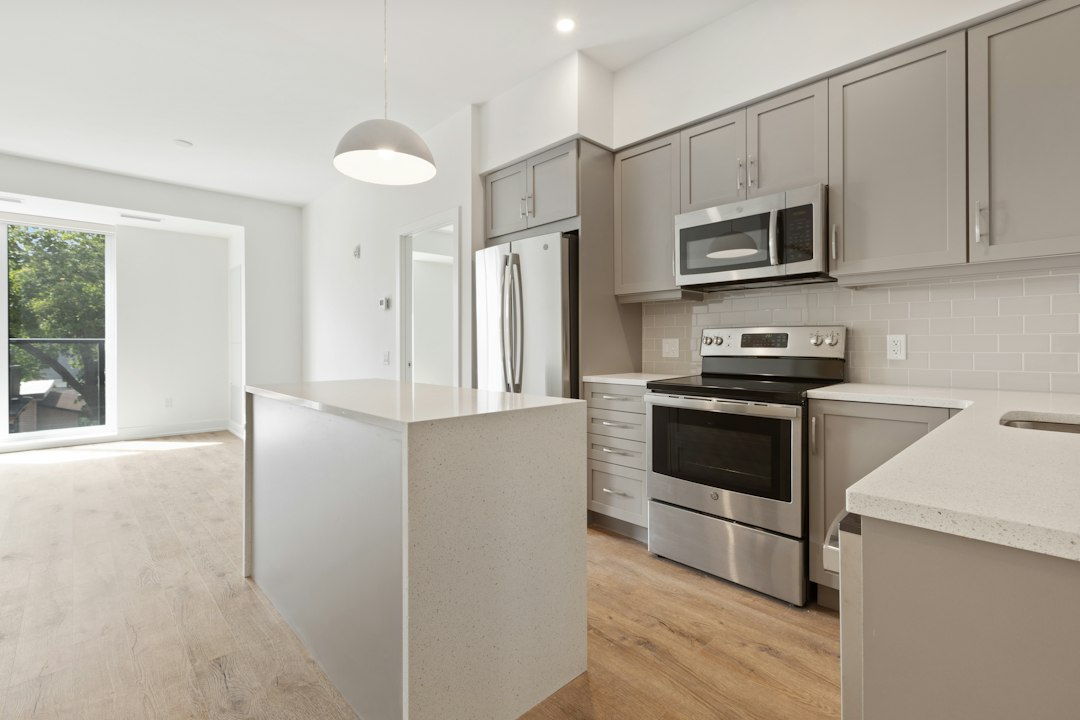 Empty renovated apartment kitchen with gray shaker cabinets, a white quartz island and counters, stainless refrigerator, range and microwave, and light oak vinyl plank flooring running to a window